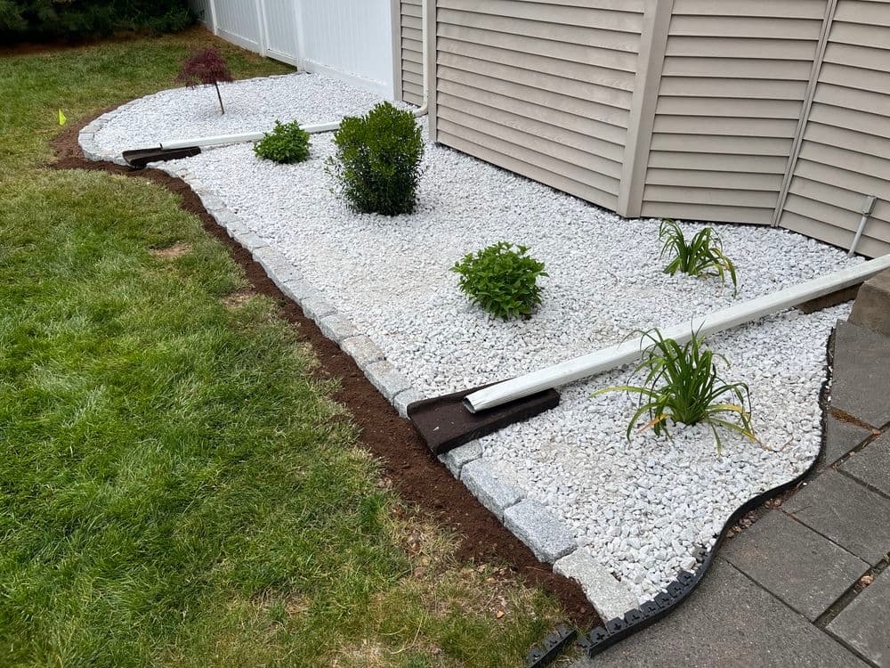 Landscaped garden with white gravel, shrubs, and drainage pipes beside a house.