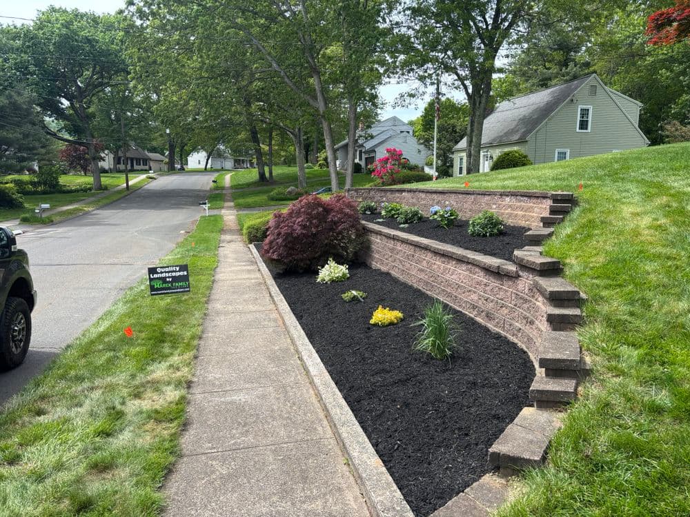 Landscaped garden with colorful plants, mulch, and stone retaining walls along a residential street.