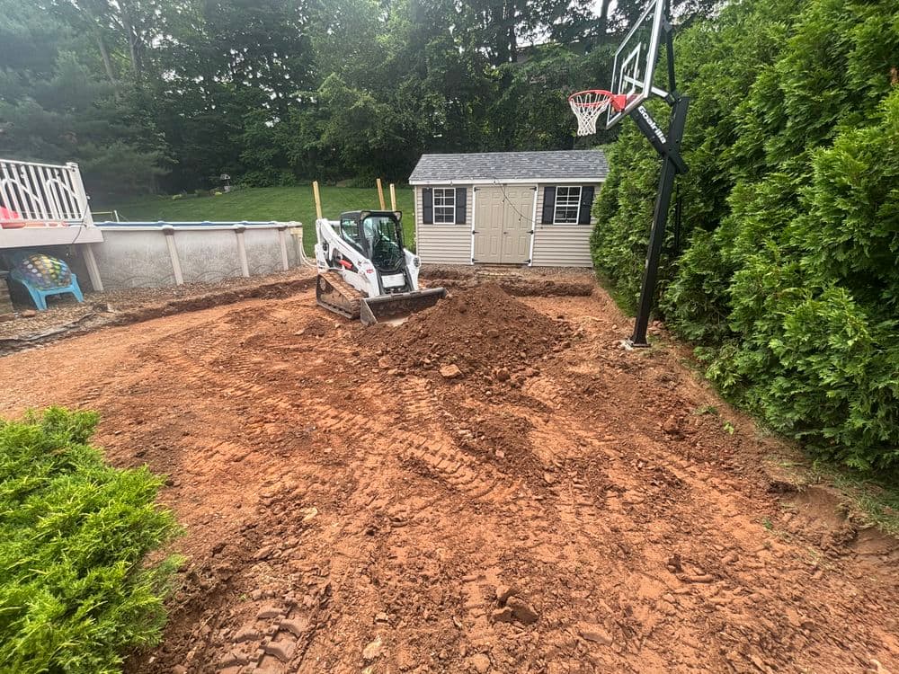 Excavator on dirt in backyard, surrounded by greenery and a basketball hoop.